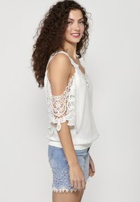 Top bianco con dettagli in pizzo sulle maniche, caratterizzato da un fit rilassato e una profonda scollatura. Abbinato a pantaloni corti di jeans azzurri con ricami floreali.