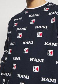Felpa blu scuro con motivo ripetuto di logo "KANI" in bianco e rosso, con scollo rotondo a costine e tessuto morbido e testurizzato.