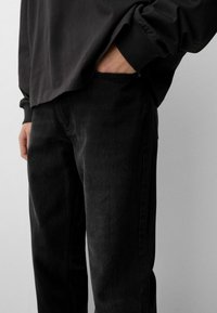 Osoba u crnim trapericama s desnom rukom u džepu i tamnom dugom rukavu na jednostavnoj svijetloj pozadini.