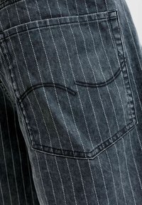Poche arrière d'un jean en denim gris foncé avec des fines rayures blanches verticales et un détail de couture noire courbée sur la poche.