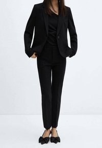 Blazer noir sur mesure et pantalon fuselé en tissu lisse, assortis à des ballerines noires ornées de nœuds. Design simple et élégant.