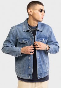 Giacca di jeans azzurro chiaro con design classico, cuciture visibili, tasche frontali e bottoni argentati, indossata sopra una maglietta nera.