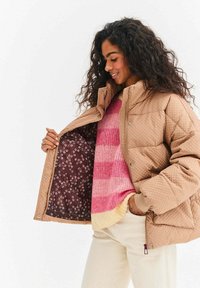 Prešívaná svetlohnedá bunda s patentnými gombíkmi, s hnedým kvetinovým podšívaním. Nosená cez ružový a krémový pruhovaný sveter a krémové nohavice.