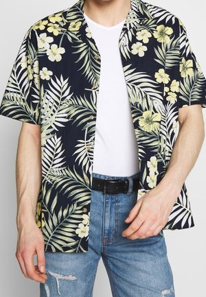 Homme portant une chemise hawaïenne noire déboutonnée avec des fleurs jaunes et des feuilles vertes sur un t-shirt blanc, un jean bleu déchiré et une ceinture noire.