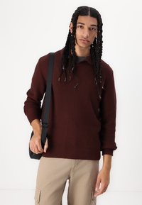 Strikket bordeaux sweater med krave og ribbet tekstur, båret med beige cargobukser og en sort skuldertaske. Modellen har fletninger.