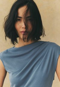 Haut drapé bleu à manches courtes, présentant une texture de tissu douce et un design asymétrique, sur un fond neutre.