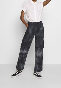 Pantalones cargo de tela oscura tie-dye, con múltiples bolsillos, un corte relajado y una altura media, combinados con una camisa blanca de botones y zapatillas negras.