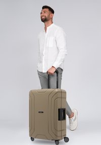 Uomo in camicia bianca e pantaloni grigi che tiene il manico di una grande valigia beige con ruote, sorridendo e guardando in alto.
