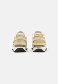 Saucony sneakers met een beige bovenwerk en mesh accenten, gele hieltab en zwarte rubberen zool. De achterkant benadrukt de branding.