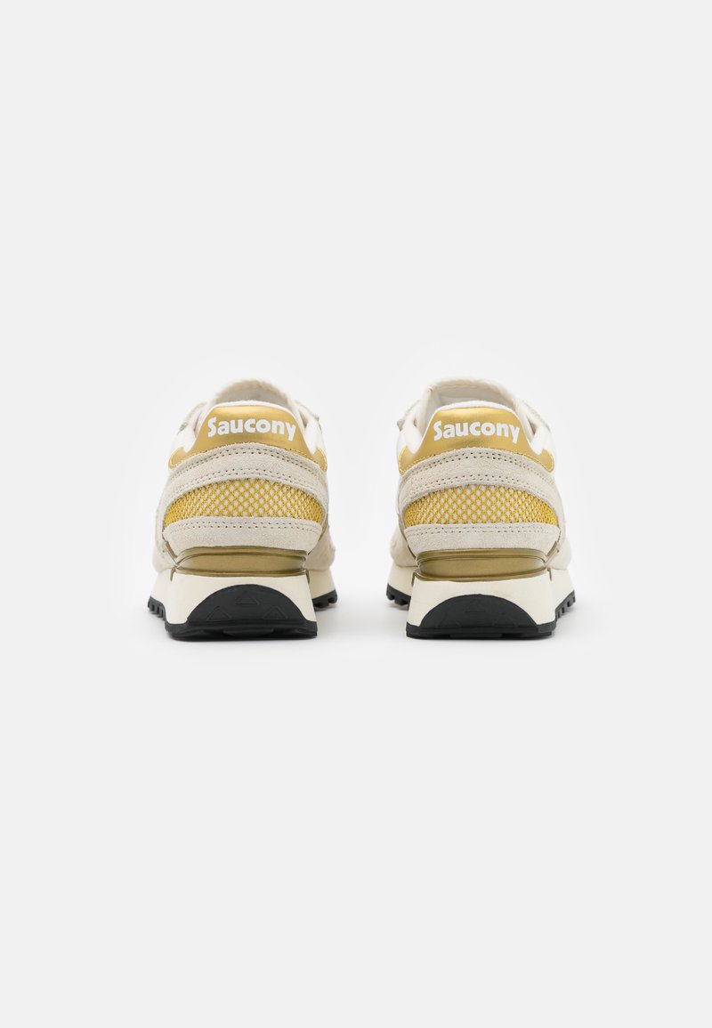 Sneakers Saucony con una parte superiore beige con accenti in mesh, un tab sul tallone giallo e una suola in gomma nera. La vista posteriore mette in evidenza il marchio.