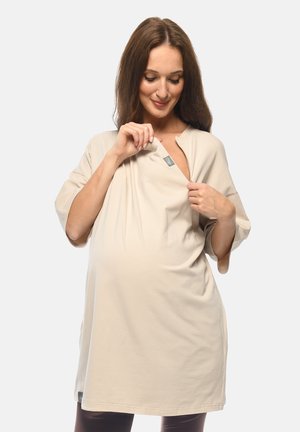Donna incinta con capelli castani lunghi che indossa una maglia premaman beige, tirando lo scollo per mostrare una piccola apertura per l'allattamento.
