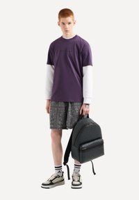 Zaino nero con texture e tasca frontale, indossato sopra pantaloni corti a fantasia grigia e una maglietta viola sovrapposta a una maglia bianca a maniche lunghe.