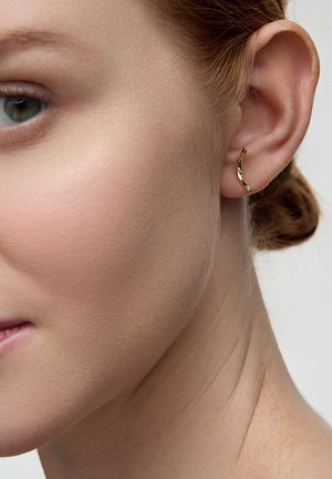 Złota kolczyk w kształcie obręczy z zakręconym wzorem, spoczywający na małżowinie usznej, prezentujący błyszczącą powierzchnię i gładką teksturę. Widoczne subtelne odbicia światła.