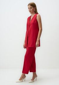 Red sleeveless blazer with a V-neck and button closure, paired with matching tailored pants. Glossy fabric and straight leg design.