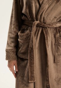 Brązowy pluszowy szlafrok o miękkiej fakturze, z wiązanym paskiem w talii, dwoma przednimi kieszeniami i prążkowanymi mankietami na rękawach.