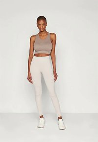 Stanik sportowy w jasnobrązowym kolorze, zestawiony z wysokimi legginsami w jasnobeżowym odcieniu. Oba elementy mają gładką fakturę oraz dopasowany krój. Całość uzupełniają białe buty sportowe.