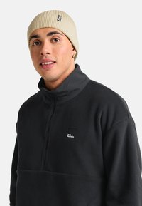 Giovane uomo che indossa un berretto di maglia beige e una giacca in pile nera Jack Wolfskin con colletto zipper, che guarda leggermente a sinistra, su uno sfondo neutro.