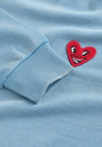Lyseblå sweatshirtærme med ribbet manchet og et rødt hjerte-patch med et smilende ansigt med øjne og tænder broderet på stoffet.