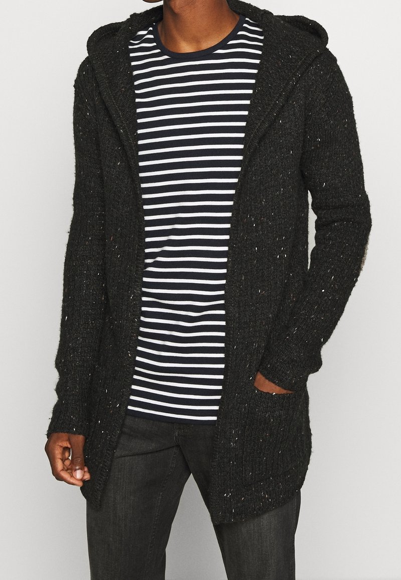Homme portant un cardigan à capuche noir moucheté par-dessus un t-shirt rayé noir et blanc avec un jean foncé, main dans la poche, sur un fond uni.