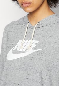 Grå huvtröja med prickig textur, vit Nike-logotyp och dragsko med tofsar vid halsen. Avslappnad, tröja att dra över huvudet.