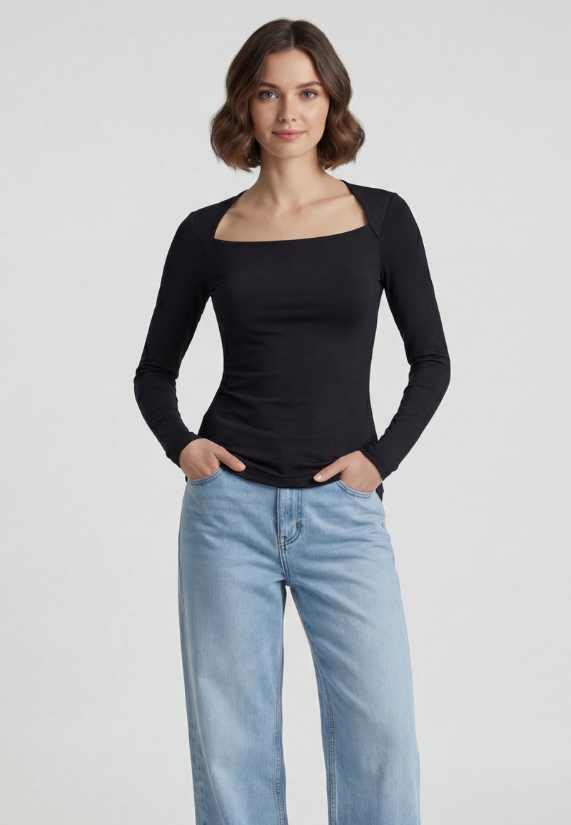 Femme aux cheveux courts et bruns portant un haut noir à manches longues avec un col carré et un jean bleu clair taille haute, mains dans les poches.