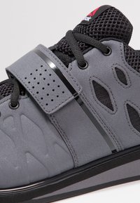 Gray athletic shoe featuring a textured synthetic upper, mesh panels, black laces, a Velcro strap, and contrasting glossy black accents.