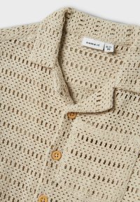 Maglia beige lavorata a maglia con texture a trama aperta, colletto corto e tre bottoni in legno sulla parte anteriore. Inclusa una piccola tasca sul petto.