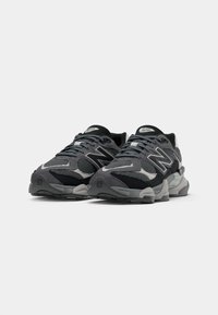 Chaussures de sport grises et noires en matériaux maille et daim, présentant un design superposé distinctif et des accents en matériel réfléchissant.