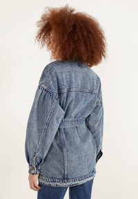 Denim jacket featuring a belted waist, balloon sleeves, and a faded blue wash. Notable for its distressed hem and classic stitching details.