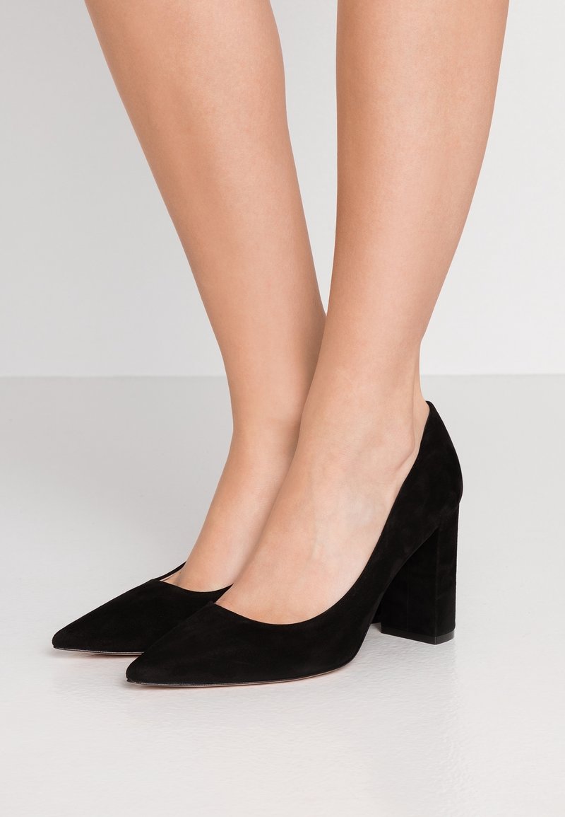 Paire d'escarpins en daim noir à bouts pointus et talons blocs portés pieds nus sur un fond uni clair.