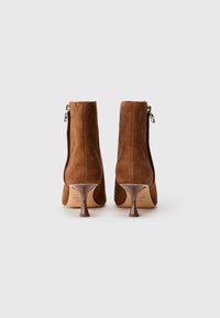Botas vaqueras de ante marrón que cuentan con un cierre de cremallera, un tacón afilado y un forro interior suave. El tacón tiene un diseño bulboso único.