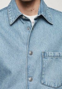 Giacca in denim azzurro chiaro con colletto classico, bottoni a pressione argentati e tasca sul petto. Il tessuto ha una consistenza morbida e cuciture visibili.