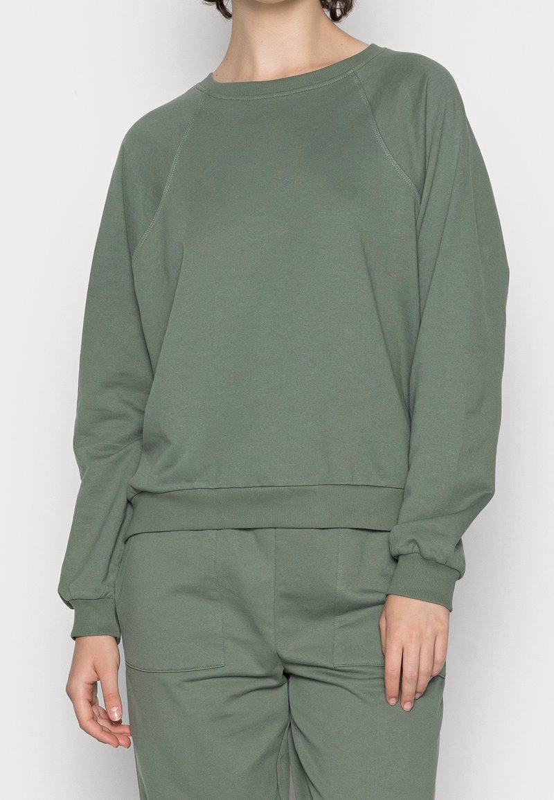 Personne portant un sweat-shirt à manches longues vert olive et un pantalon assorti, debout devant un fond uni clair.