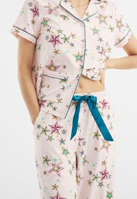 Conjunto de pijama rosa claro con estampado de estrellas en verde, púrpura y azul. Camiseta de manga corta con bolsillo; los pantalones tienen cordón de color teal.