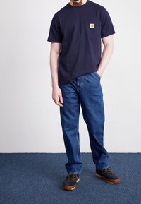Camiseta de algodón azul marino con bolsillo en el pecho y logo, combinada con jeans de mezclilla azules y zapatos oscuros de cordones con suela color tan.