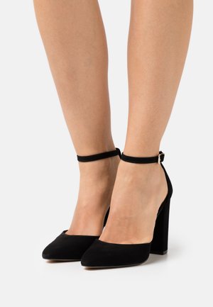 Black suede high-heeled pumps with a pointed toe and ankle strap, featuring a chunky heel for stability.