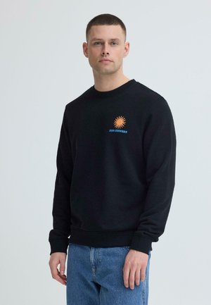 Man met een zwart sweatshirt met oranje zonlogo en de tekst "SUN SEEKERS", gecombineerd met een blauwe spijkerbroek, staand tegen een effen achtergrond.