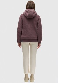 Haut en molleton de couleur violet pâle, avec une surface douce et texturée, une capuche avec cordon de serrage et un ourlet côtelé. Porté avec un pantalon crème.