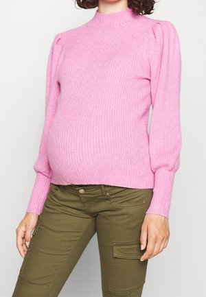 Kobieta ubrana w prążkowany różowy sweter z długim rękawem i oliwkowe spodnie typu cargo, pozująca ze swobodnie opuszczonymi rękami.