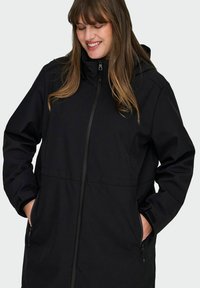 Chaqueta negra con capucha, cierre frontal y bolsillos laterales, con un corte relajado. Presenta una textura suave y sin patrones o acentos visibles.
