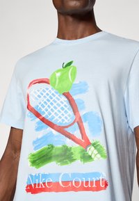 Svetlomodré bavlnené tričko s grafickým dizajnom červenej tenisovej rakety so zeleným jablkom a textom "Nike Court" v červenej a modrej farbe.