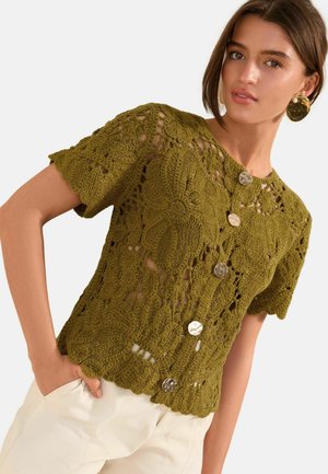 Frau trägt olivgrüne, gehäkelte kurzärmelige Strickjacke mit großen Knöpfen und gewellten Rändern, kombiniert mit hellbeigen Hosen.