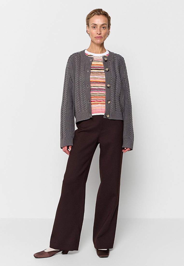 Cardigan en maille torsadée grise avec un col rond et une fermeture à boutons, superposé à une chemise rayée. Associé à un pantalon large marron et des chaussures sombres.