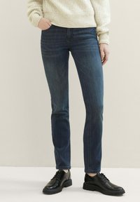 Jeans de mezclilla azul oscuro con un ajuste slim y sutilezas de desgaste. Combinados con unos zapatos de cordones negros brillantes y un suéter texturizado crema.