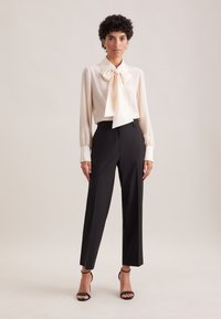 Cream blouse with a large bow tie detail, long sleeves, and structured cuffs; paired with black tailored trousers and black strappy heels.