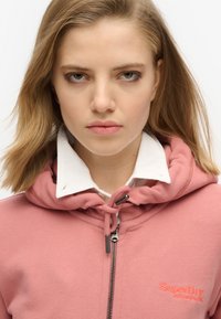 Joven con cabello rubio que lleva una sudadera rosa con cremallera sobre una camisa blanca con cuello, mirando directamente al frente con una expresión neutral.