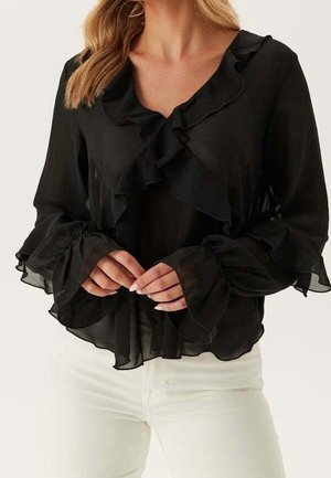 Zwarte doorzichtige blouse met gerimpelde kraag, voorkant en uitlopende manchetten, gedragen met een witte broek, handen op de taille gevouwen.