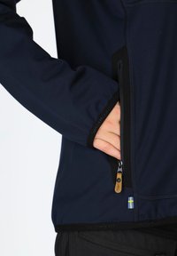 Navy Softshell-Jacke mit einer Reißverschlusstasche mit Lederzieher und schwedischem Flaggenakzent. Schwarze Abschlüsse an den Bündchen und dem Saum.