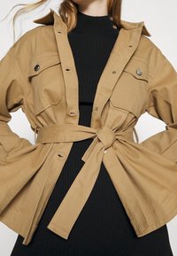 Veste beige ceinturée avec poches poitrine à boutons et manches larges, superposée sur une robe noire à côtes. Fabriquée dans un tissu structuré et durable.