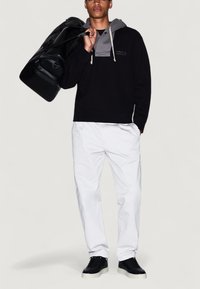 Hombre con sudadera negra y gris, pantalones blancos, zapatillas negras, llevando una bolsa de deporte negra sobre el hombro frente a un fondo liso.
